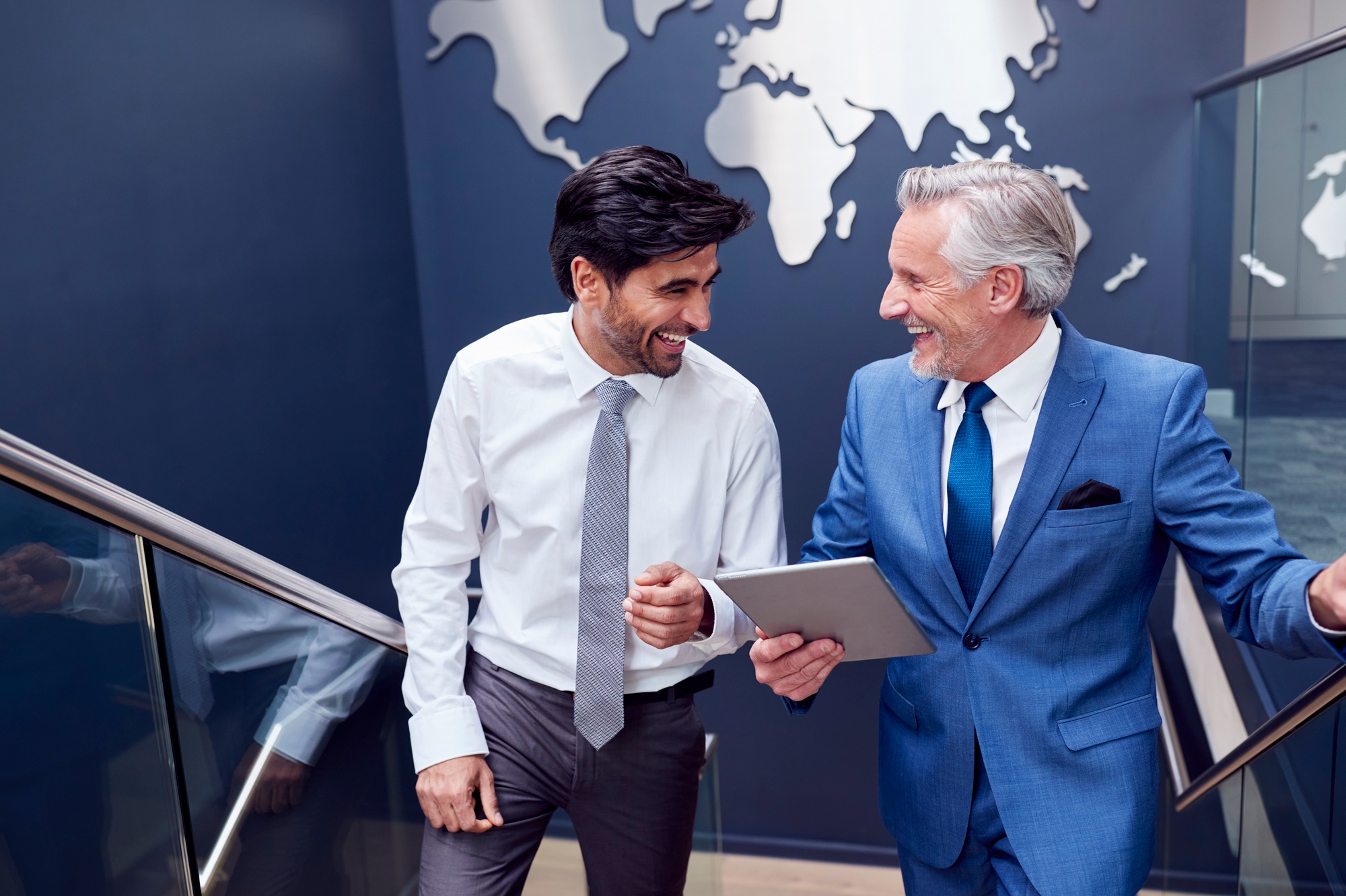 Male Colleagues With Digital Tablet Meeting On Stairs Of Office With World Map In Background