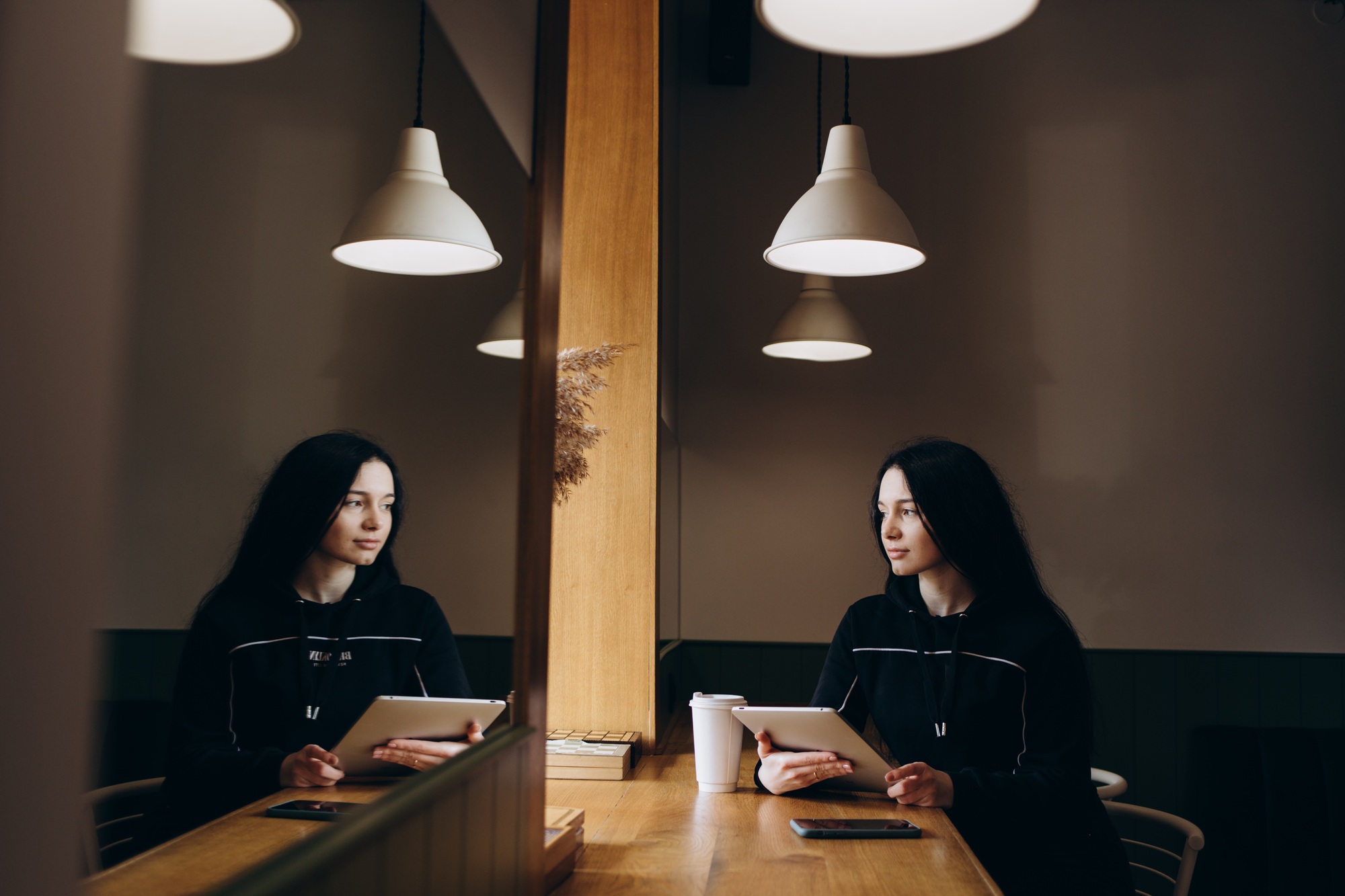 Young woman sitting in coffee shop at wooden table, drinking coffee and using smartphone.